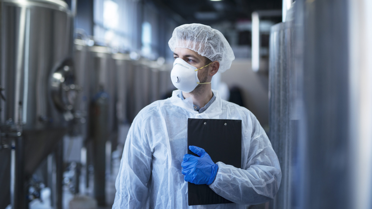 Technologist in protective white suit with hairnet and mask standing in food factory.